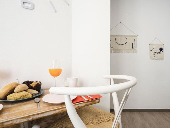 A dining area with a wooden table, white chairs, a basket, a plate of bread, a glass of orange juice, and a white mug.