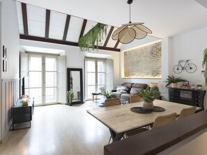 A spacious living area with a high ceiling, large windows, a dining table, and a sofa.