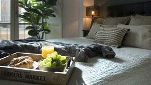 Una cama bien hecha con un edredón gris, una lámpara de noche y una ventana con vistas a edificios.