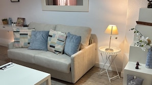 A living room with a sofa, a side table with a lamp, a small table, and a decorative pillow. - Santelmo 16 (Málaga)