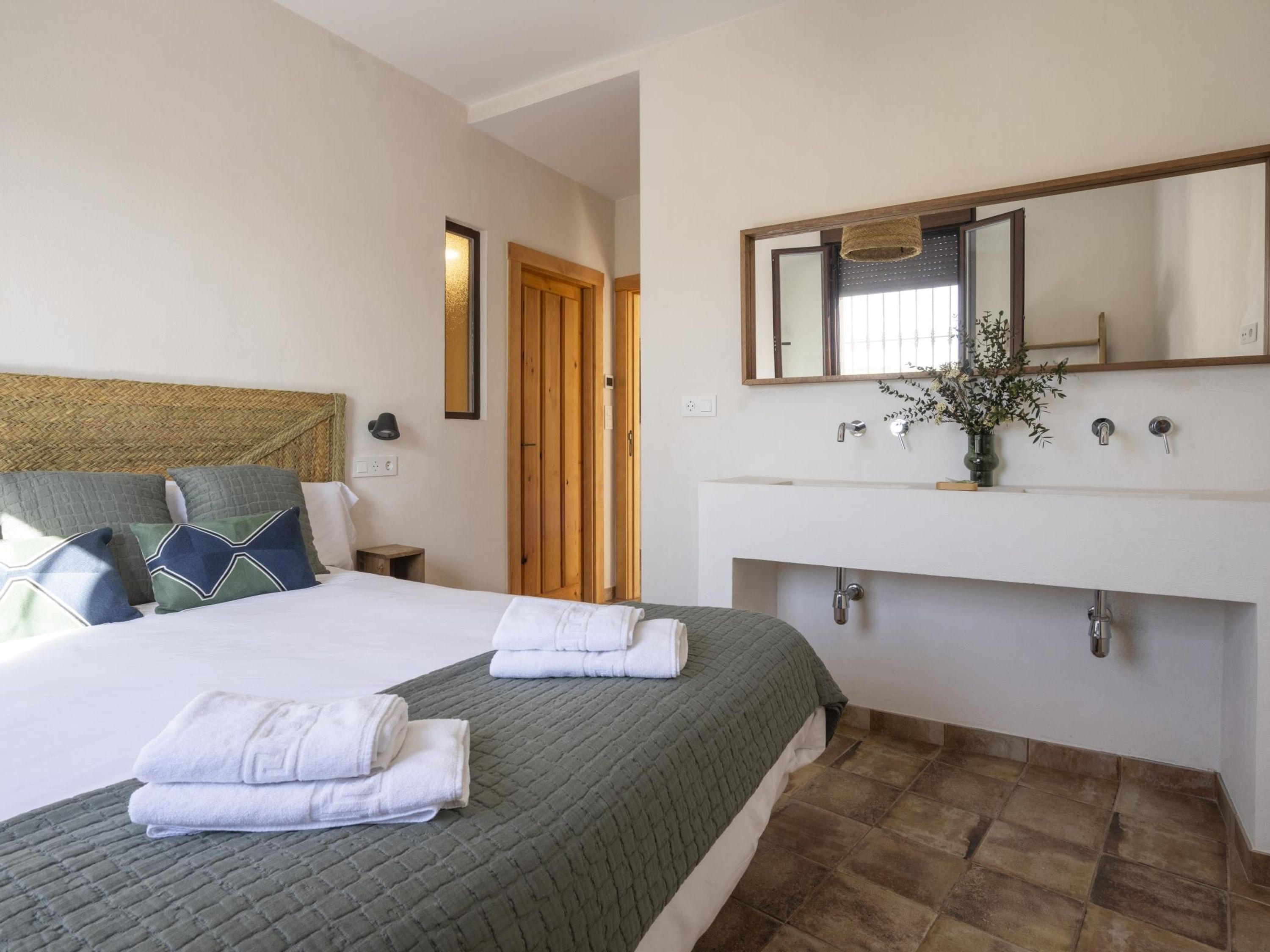 A hotel room with a bed, a bedside table, a nightstand, a large mirror, a white sink, and a door leading to another room.