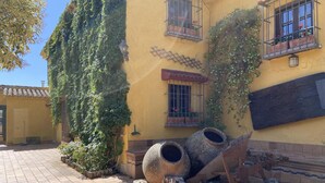 A courtyard with tiled flooring, large stone planters, and a yellow building with climbing plants and barred windows. - Casa Rural del Jardin (Priego de Córdoba)