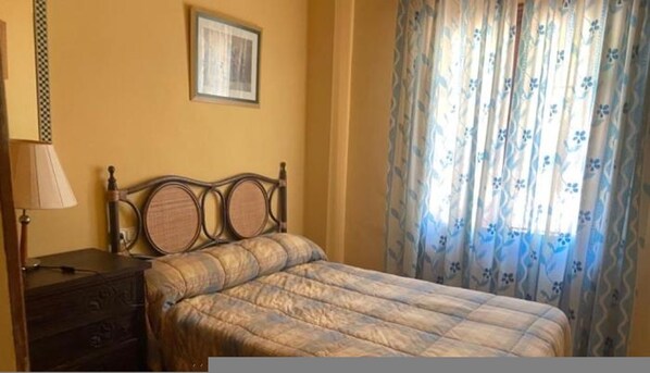 Un dormitorio con cama, mesita de noche, lámpara, un cuadro enmarcado en la pared y una ventana con cortinas florales.
