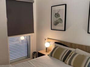 Une chambre à coucher avec un lit, une lampe de chevet, une fenêtre avec des stores et une plante décorative accrochée au mur.