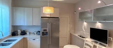 A compact kitchen with a Bosch refrigerator, a dishwasher, a coffee maker, and a microwave. There's a dining area with a table and chairs, and a flat-screen TV mounted on the wall.