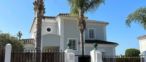 A two-story house with a white facade, a black gate, and a palm tree in the foreground.