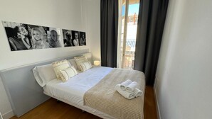 Ein ordentlich bezogenes Bett mit weißer Decke und Kissen, ein kleiner Nachttisch mit Lampe sowie ein Balkon mit dunklen Vorhängen.
