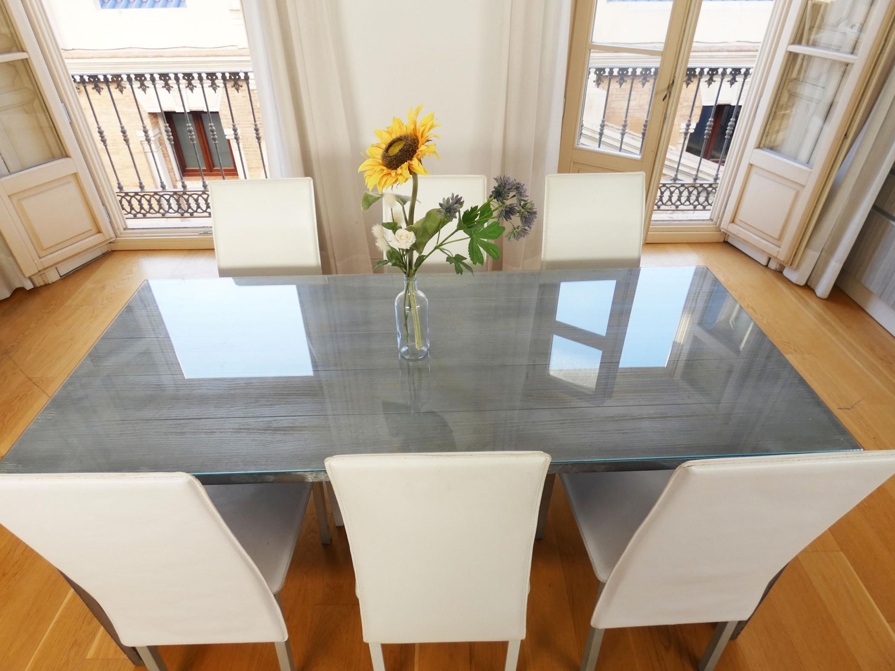 Comedor con mesa de vidrio y sillas blancas, un florero con flores y vistas a balcones.