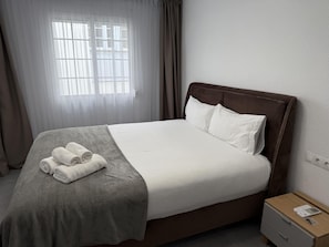 Una cama bien hecha con un edredón gris y toallas enrolladas, una mesita de noche de madera con un control remoto, y una ventana con cortinas translúcidas.
