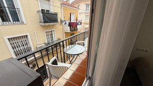 A balcony with a round glass table and two white chairs, overlooking a residential area with yellow buildings and a red-tiled roof.