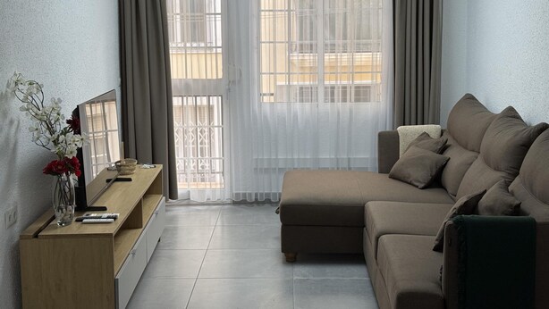 A living room with a sofa, a wooden side table, a vase with flowers, and a window with curtains.