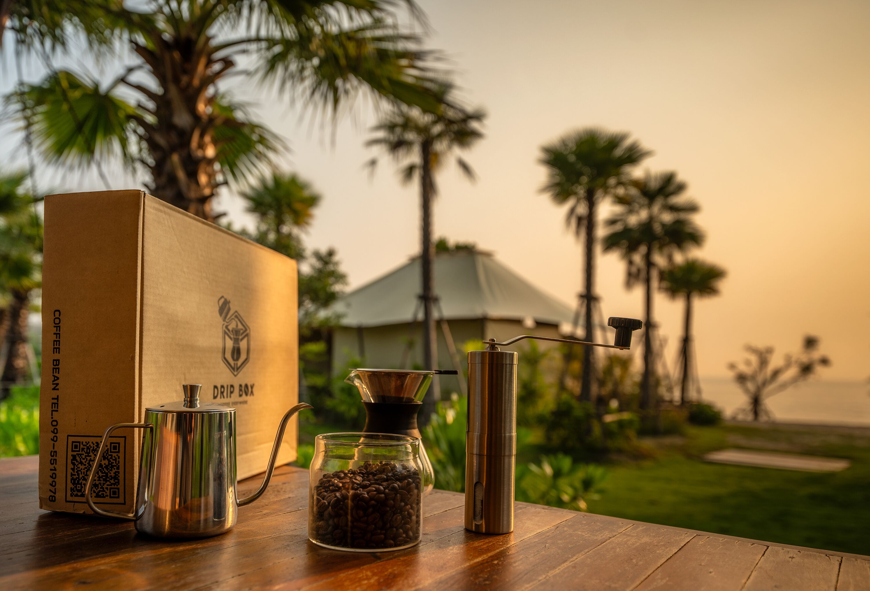 Een koffiezetapparaat, een glazen pot met koffiebonen, een koffiefilterhouder en een metalen bak op een houten tafel, met op de achtergrond palmbomen en een gebouw.