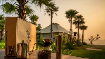 Een koffiezetapparaat, een glazen pot met koffiebonen, een koffiefilterhouder en een metalen bak op een houten tafel, met op de achtergrond palmbomen en een gebouw.