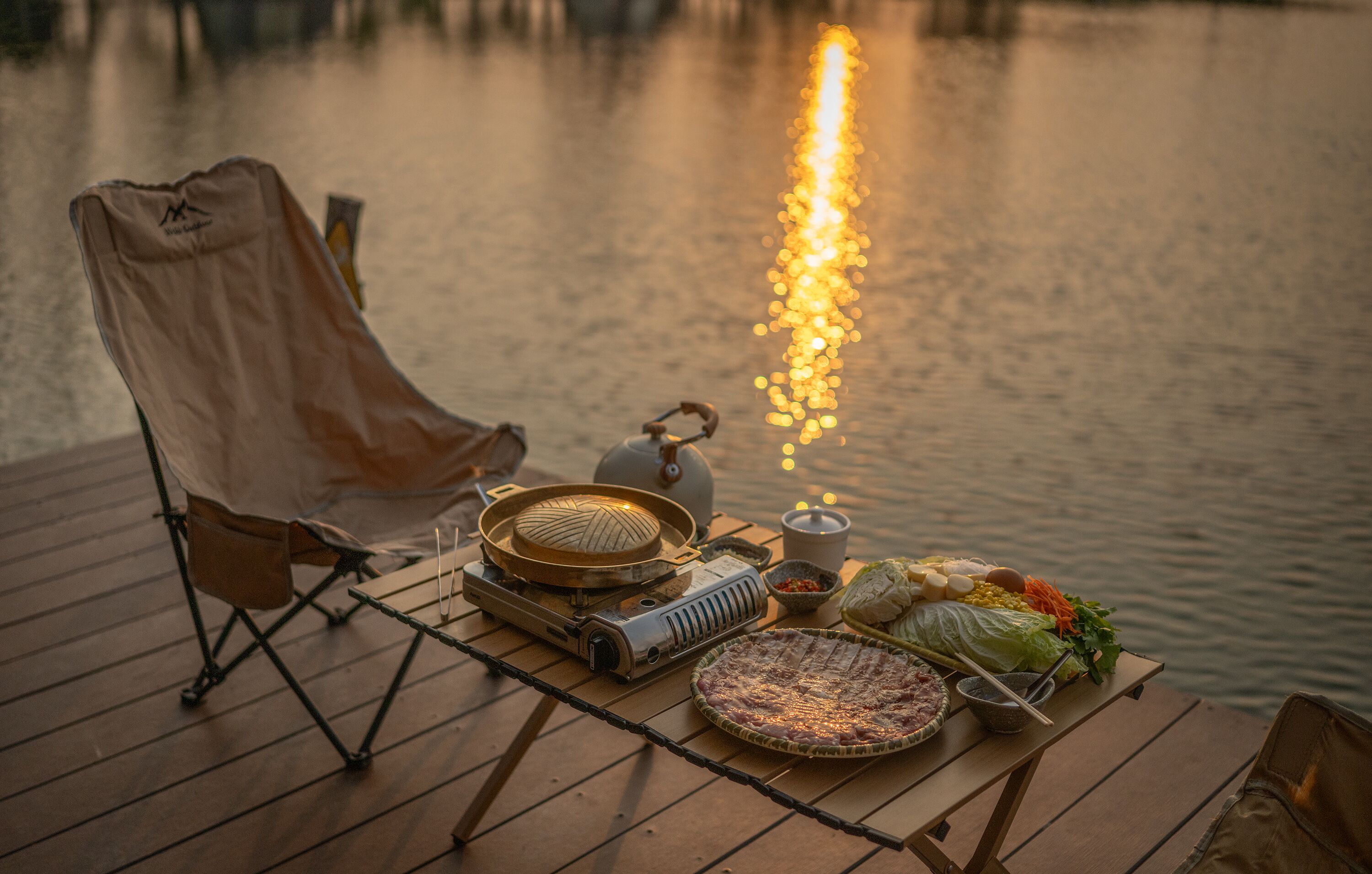 A dining setup on a wooden deck by a lake with a grill, tableware, and a chair.