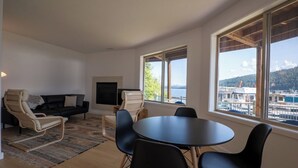 Un salon moderne avec un canapé noir, des fauteuils beiges, une table ronde et un téléviseur. De grandes fenêtres offrent une vue sur les bateaux et les collines.