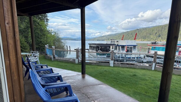 Un espace extérieur abrité, avec des chaises en plastique bleues, un sol en béton et une vue sur un quai où sont amarrés des bateaux, ainsi qu’une colline boisée en arrière-plan.