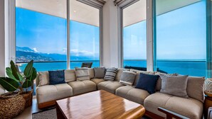 Un salon moderne doté d’une grande fenêtre offrant une vue sur la mer et le paysage urbain.