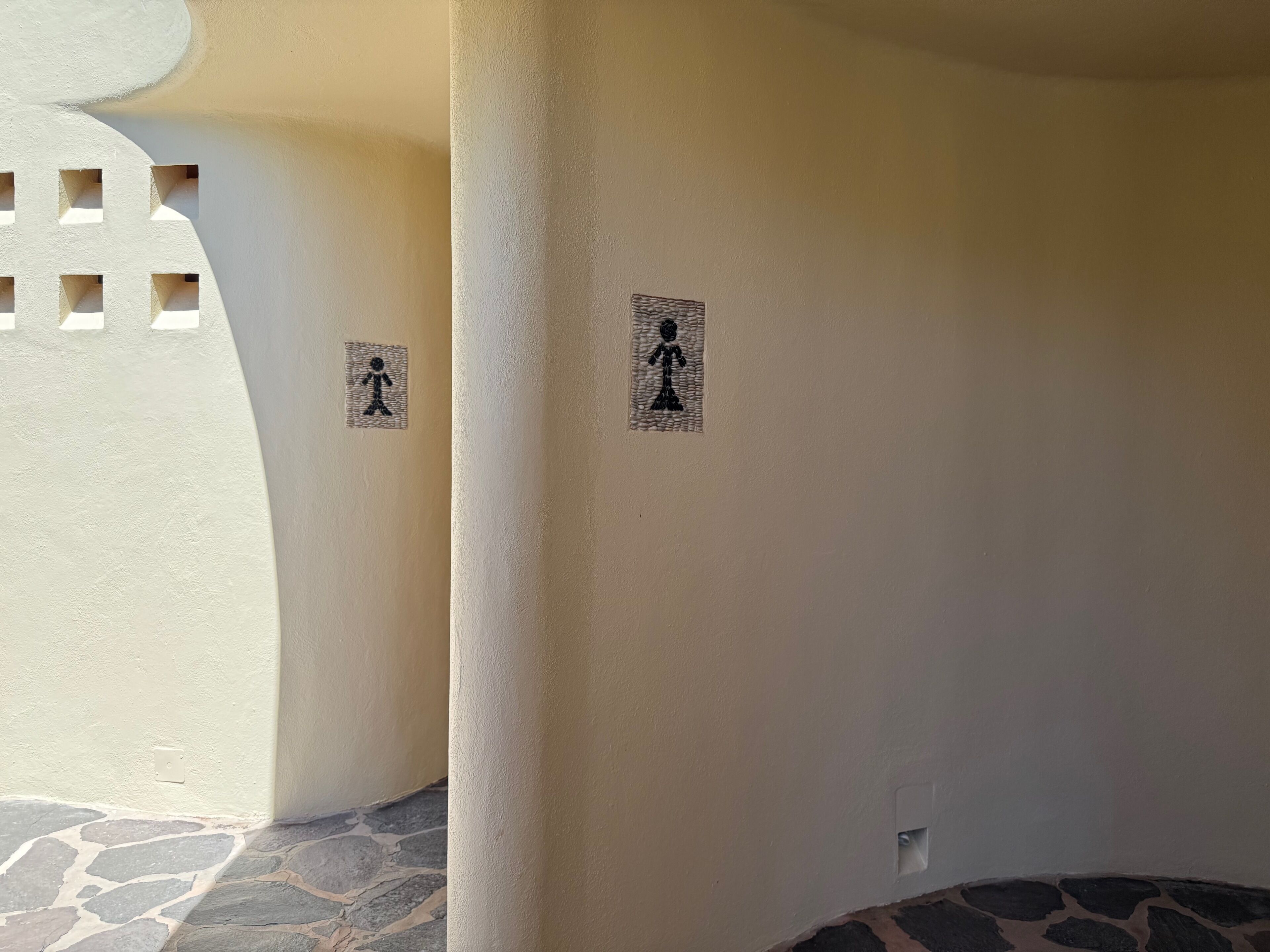 Een gang met een stenen vloertegel en muren met twee zwarte figuren die een toilet aangeven.