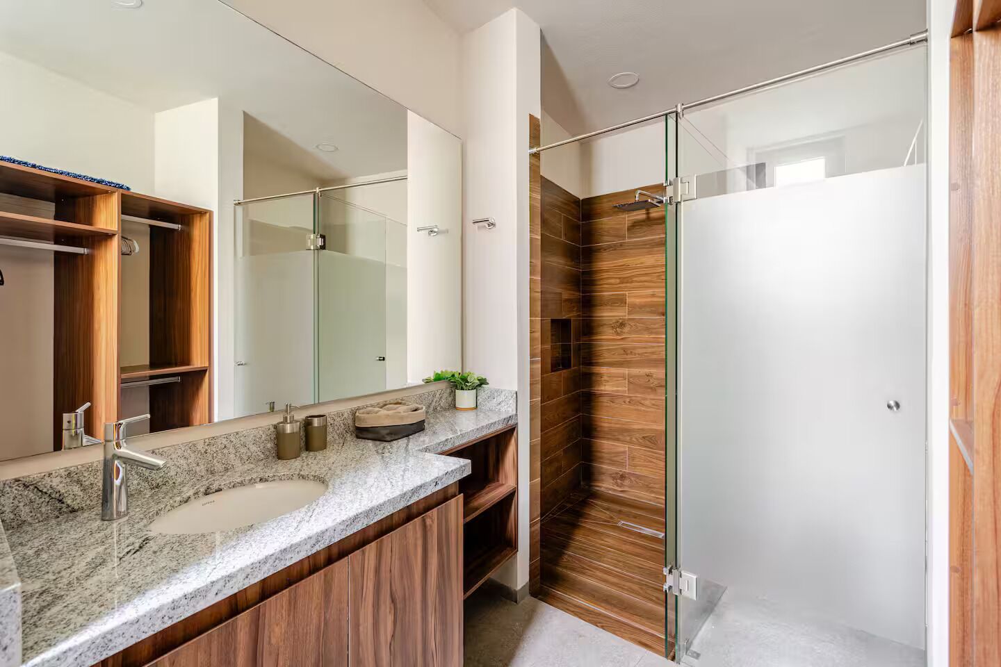 A modern bathroom with a granite countertop, wooden cabinets, a glass shower enclosure, and a large mirror.