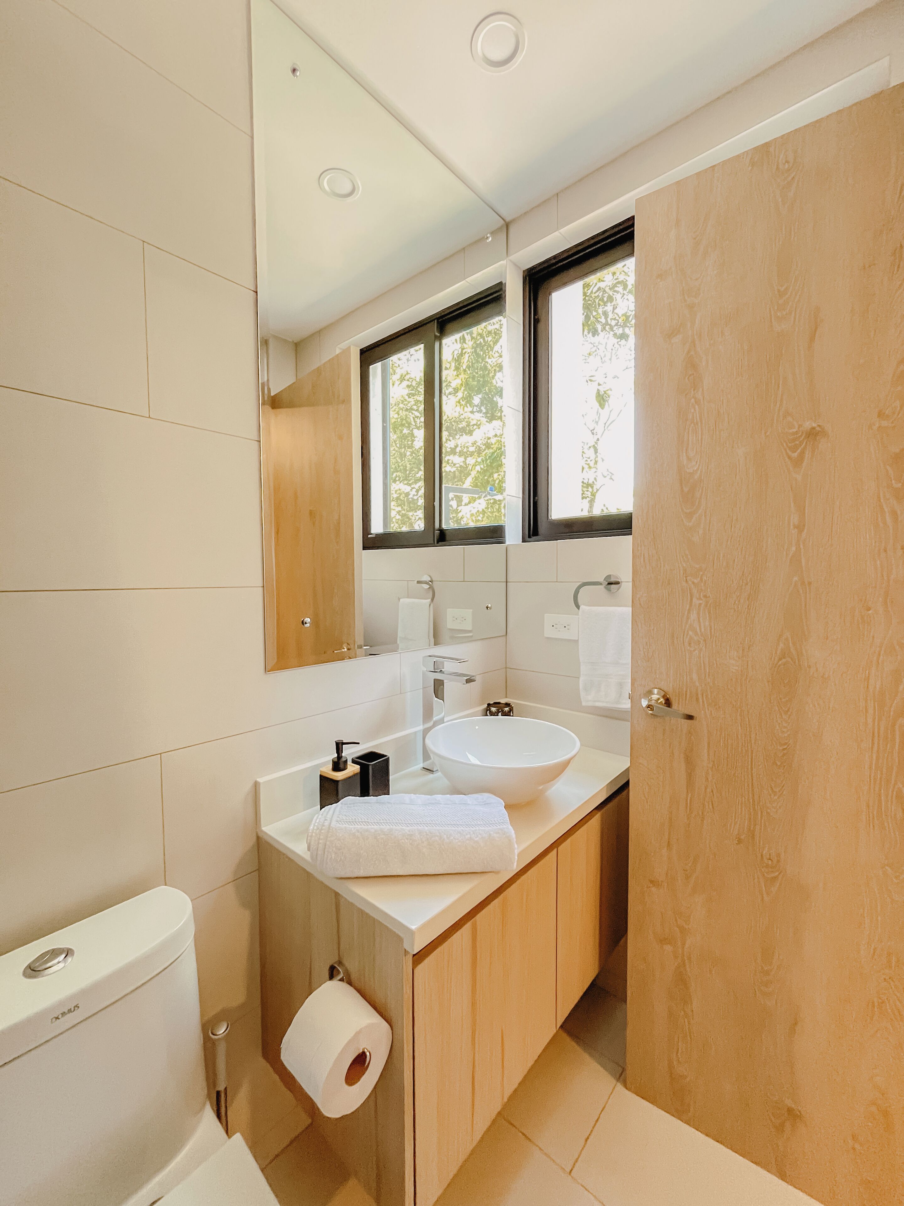 Een moderne badkamer met een witte wastafel, houten kastjes, een toilet en een raam voor natuurlijk licht.