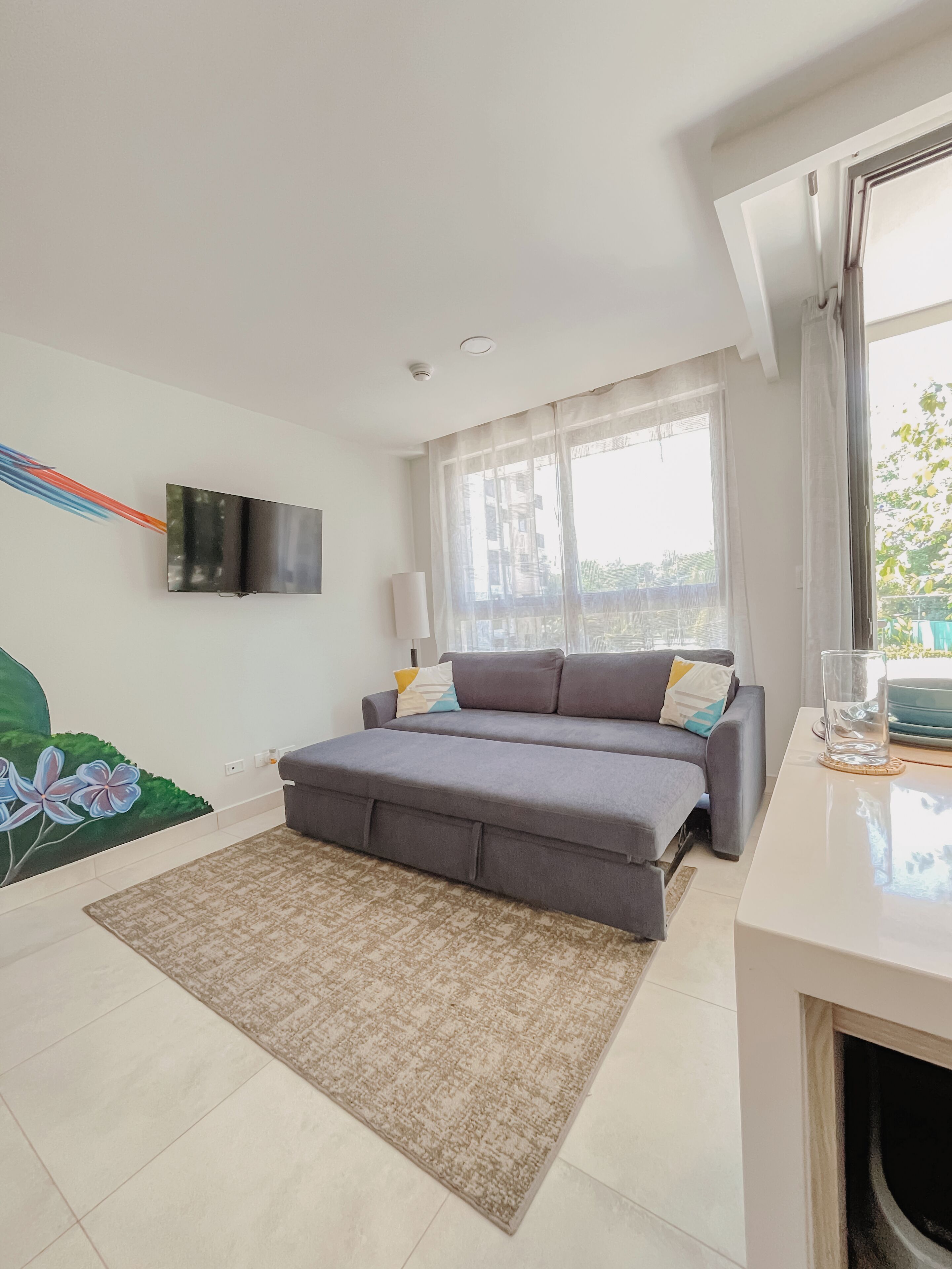 Una sala de estar moderna con un sofá gris, un televisor de pantalla plana montado en la pared, un ventanal con cortinas translúcidas y un piso de baldosas.