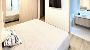 Una camera da letto con un letto, una TV fissata al muro e un bagno visibile attraverso una porta aperta.