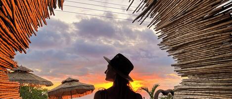 A woman sitting at a table with a view of a sunset and thatched umbrellas.