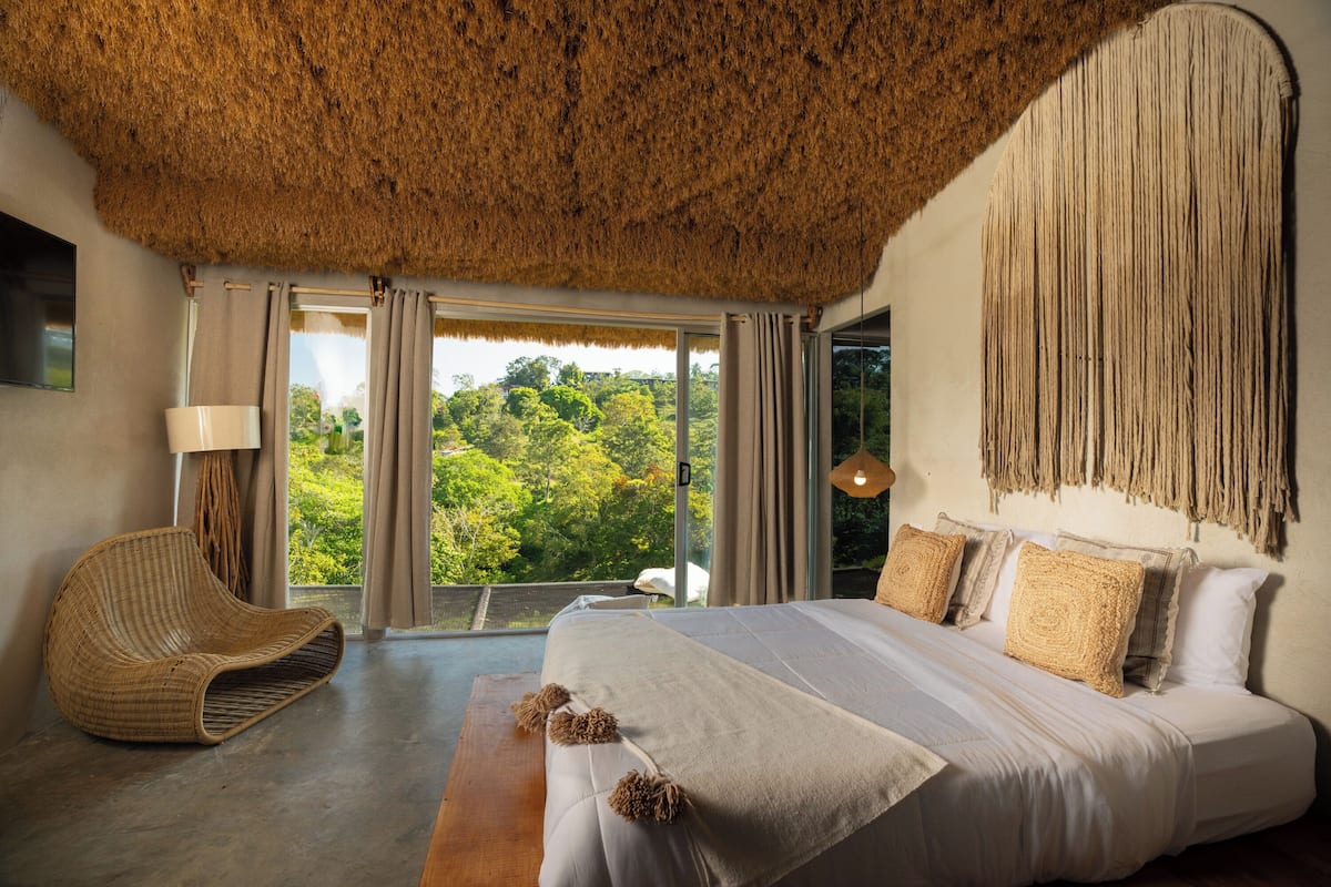 A bedroom with a large bed, a woven chair, and a view of greenery through the window.