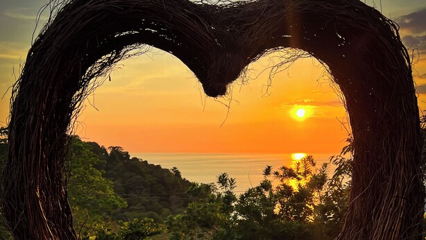 A heart-shaped wicker structure with two glasses of red liquid on a wooden surface, overlooking a sunset and landscape. - Dar Lamia Boutique Hotel (Quepos)