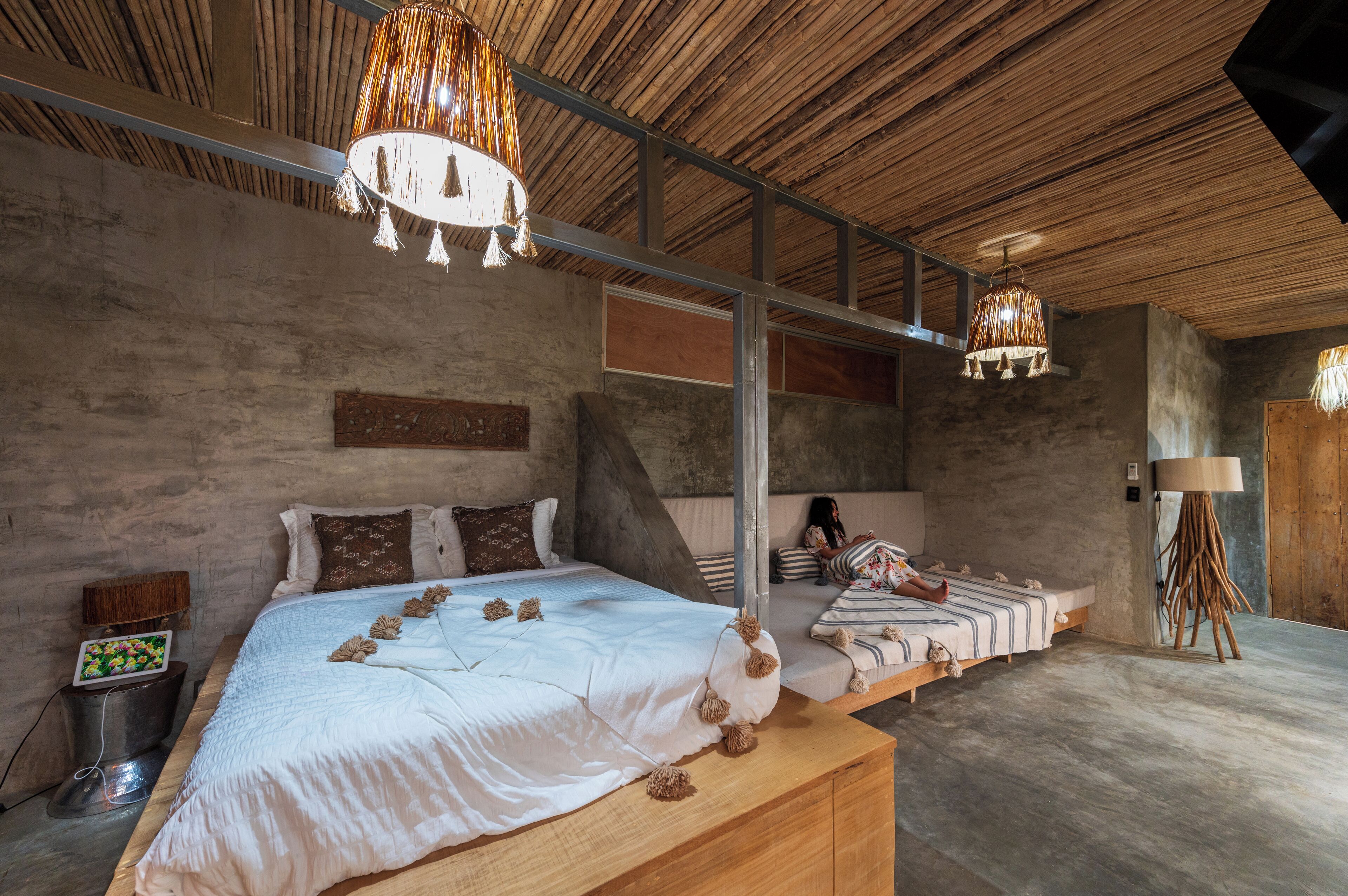Un dormitorio con una cama grande, una cama más pequeña y una mesita de noche. El techo es de madera y hay una lámpara colgante.