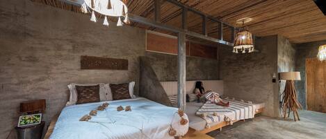 Un dormitorio con una cama grande, una cama más pequeña y una mesita de noche. El techo es de madera y hay una lámpara colgante.