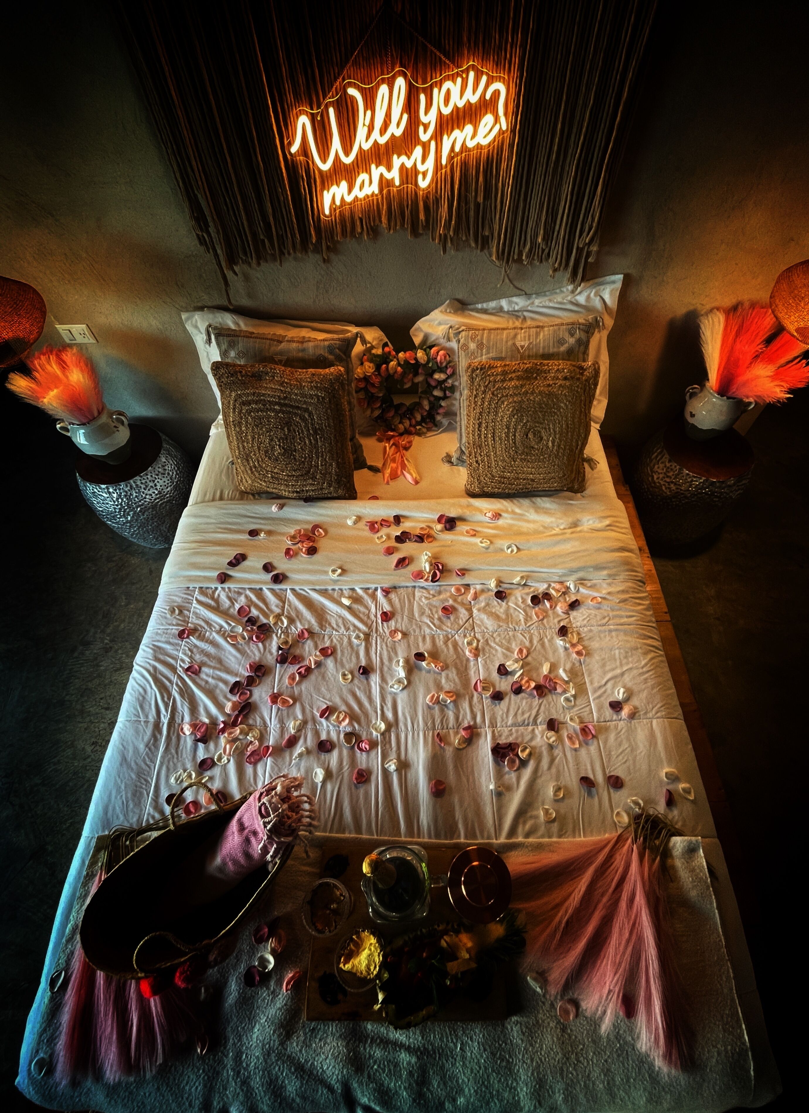 A bed with a neon sign reading "Will you marry me?" and scattered rose petals.