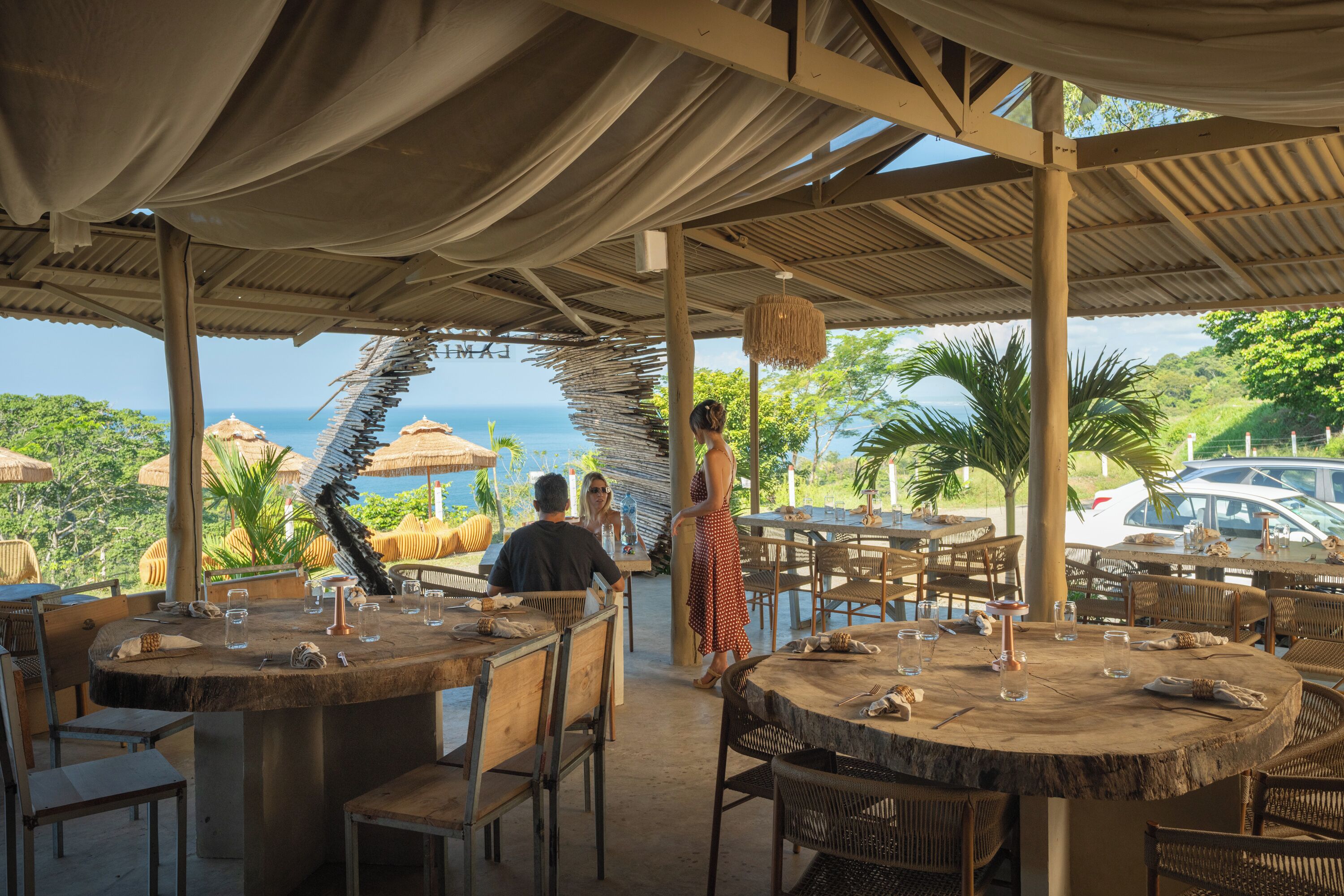 Un espacio de comedor al aire libre con mesas y sillas redondas, con vistas a un paisaje marino y vegetación exuberante.