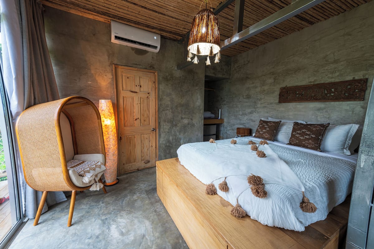 A bedroom with a large bed, a rattan chair, a hanging light fixture, and a wooden door.