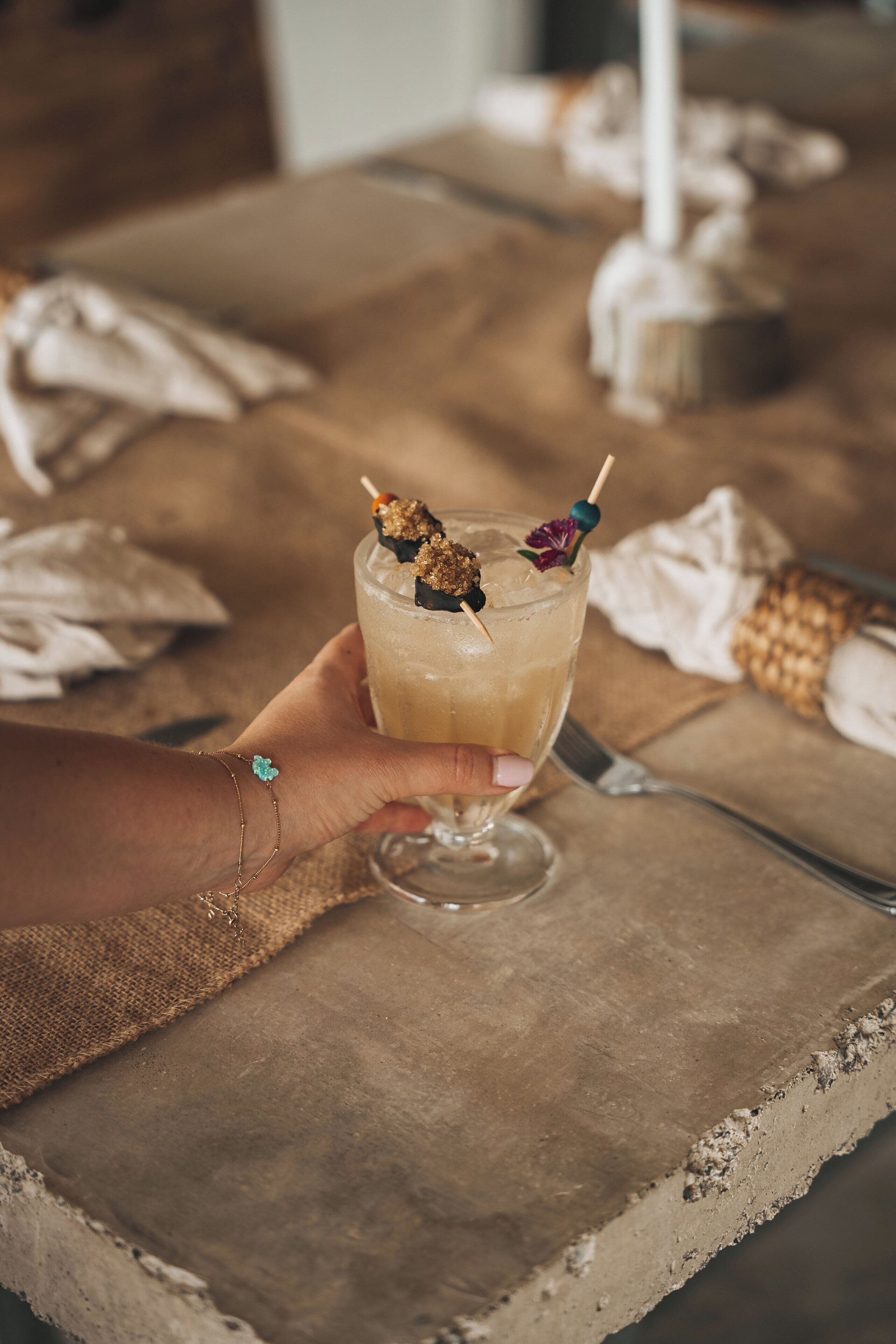 Une main tenant un verre à cocktail agrémenté d’une garniture, posé sur une table rustique avec une bougie et une serviette en tissu.