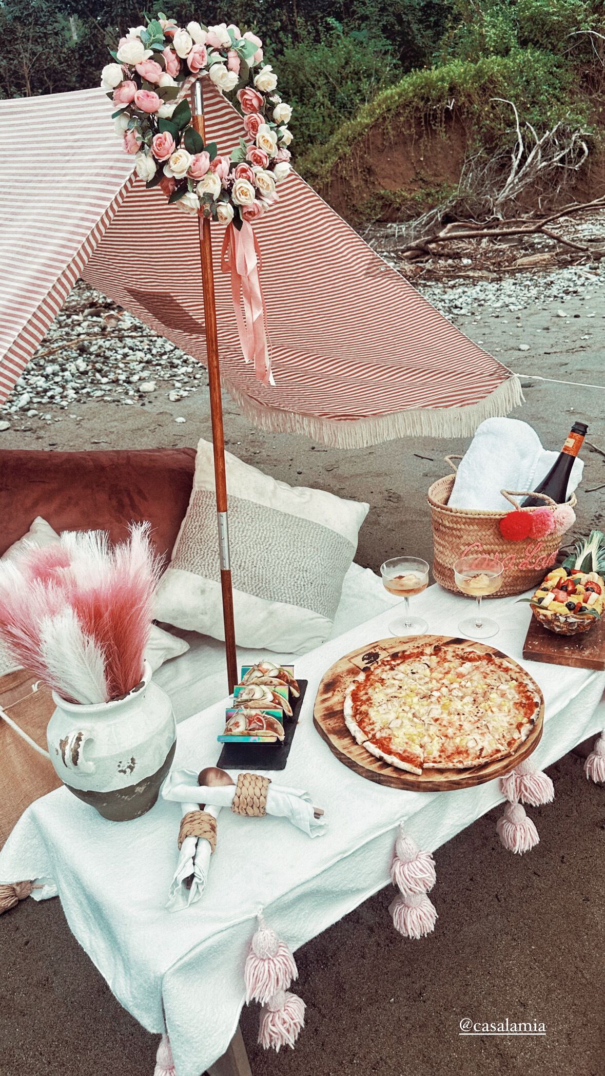 Un coin pique-nique aménagé sur la plage avec une pizza, des boissons, des en-cas et un parasol décoratif.