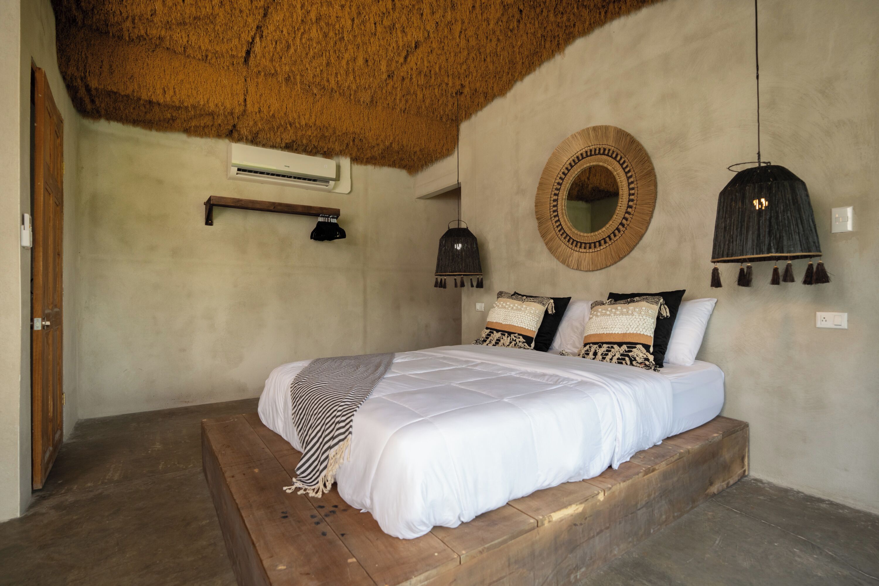 Quarto com cama, um banco de madeira, espelho redondo e luminária pendente.