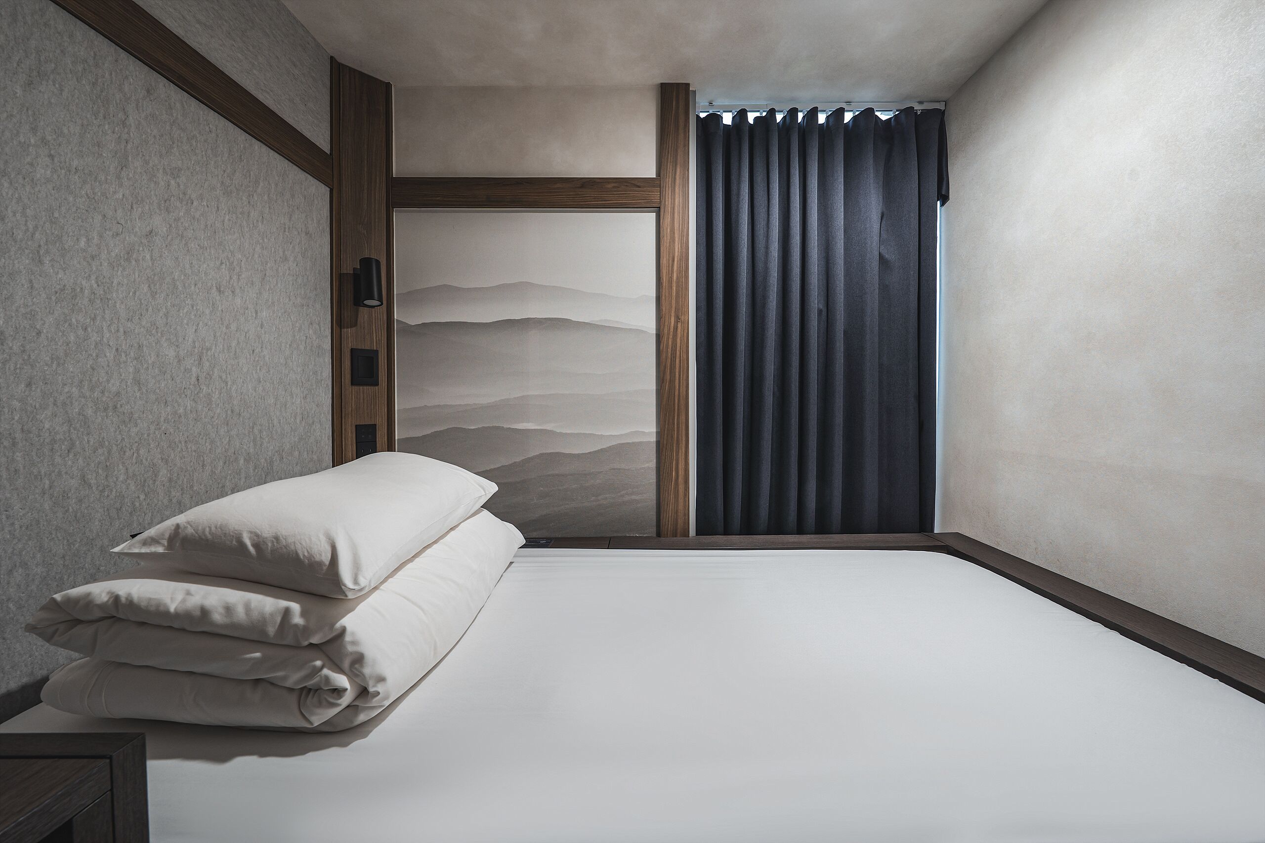 A bedroom with a bed, pillows, a wall-mounted light, and a curtain.