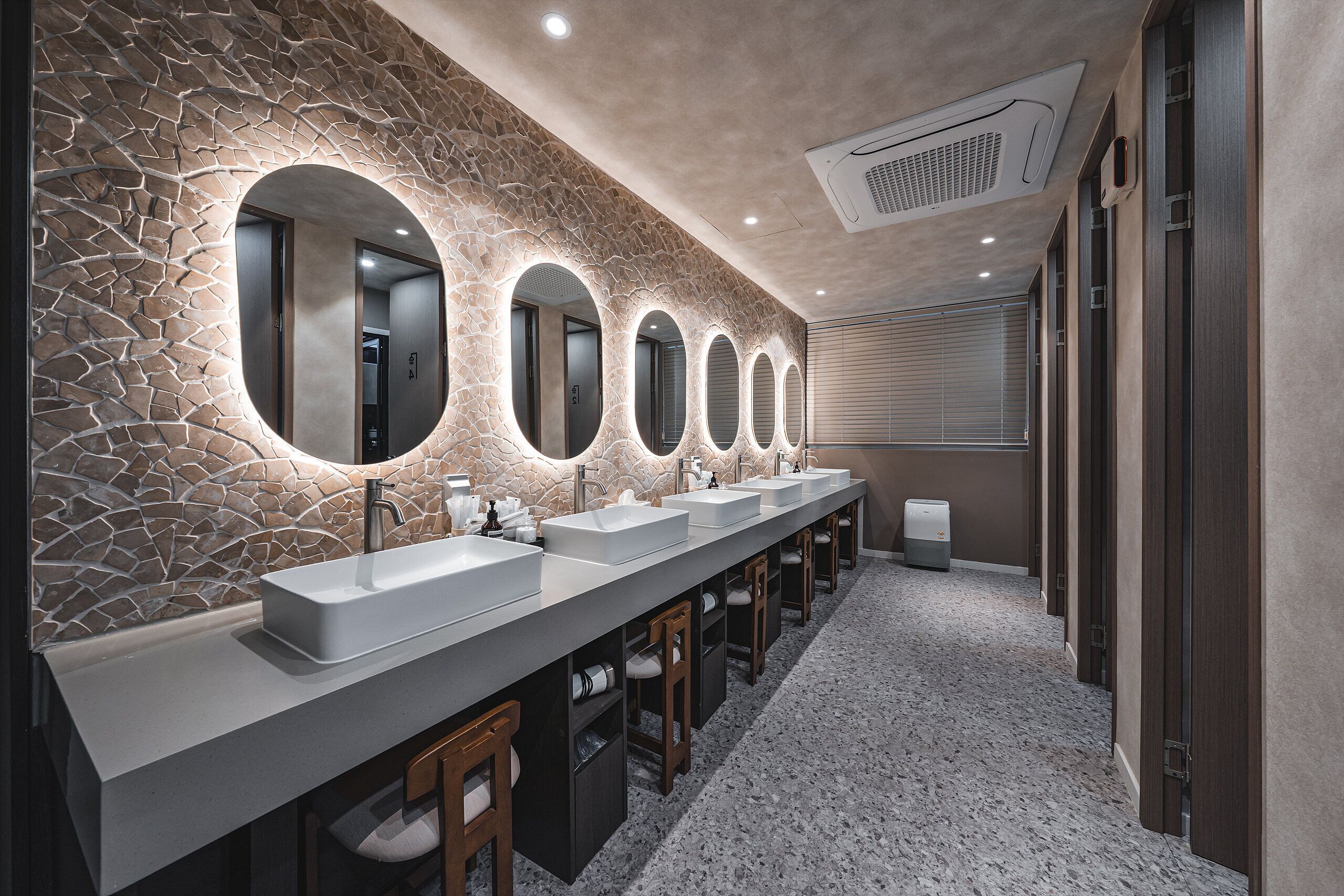 Een moderne badkamer met een stenen wand, meerdere wastafels en verlichte spiegels.
