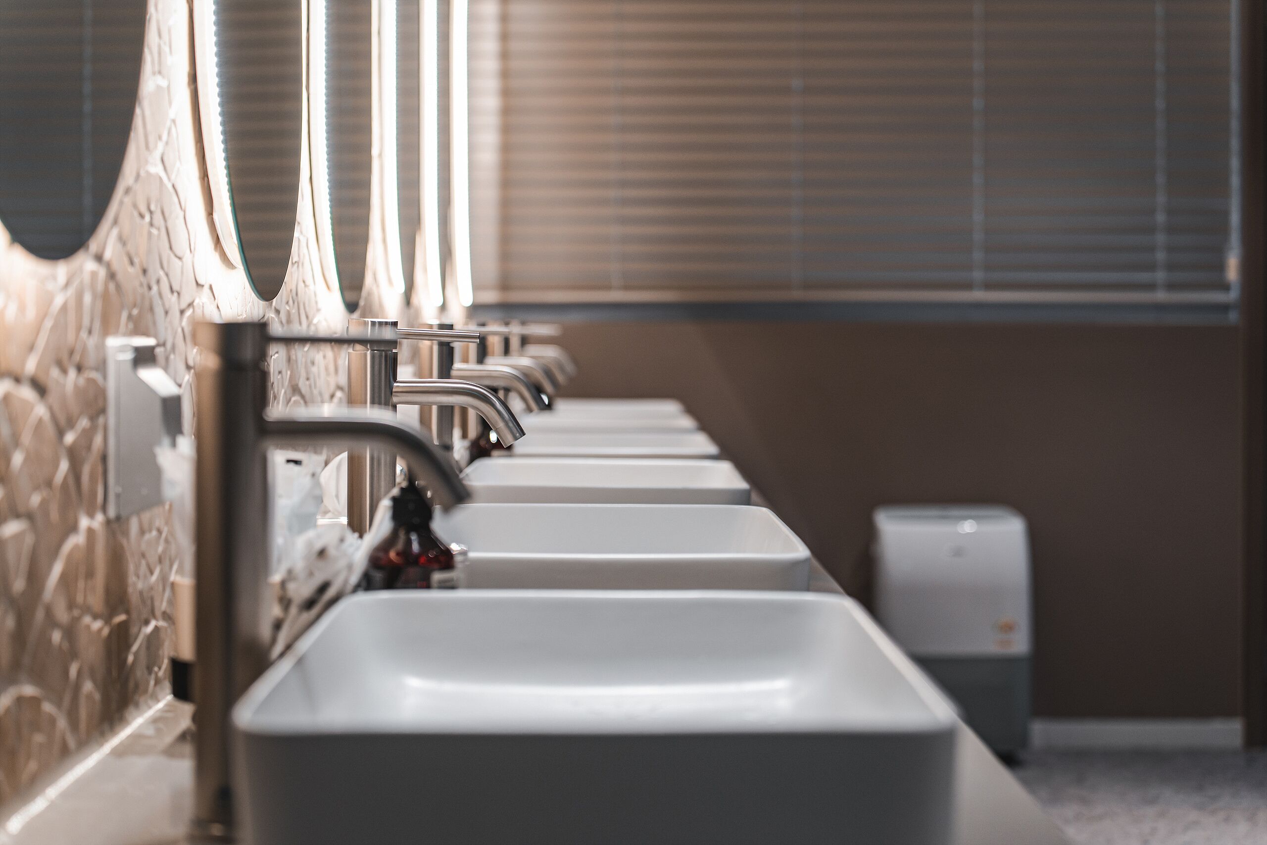 A row of sinks with a stone wall and a window with blinds in the background.