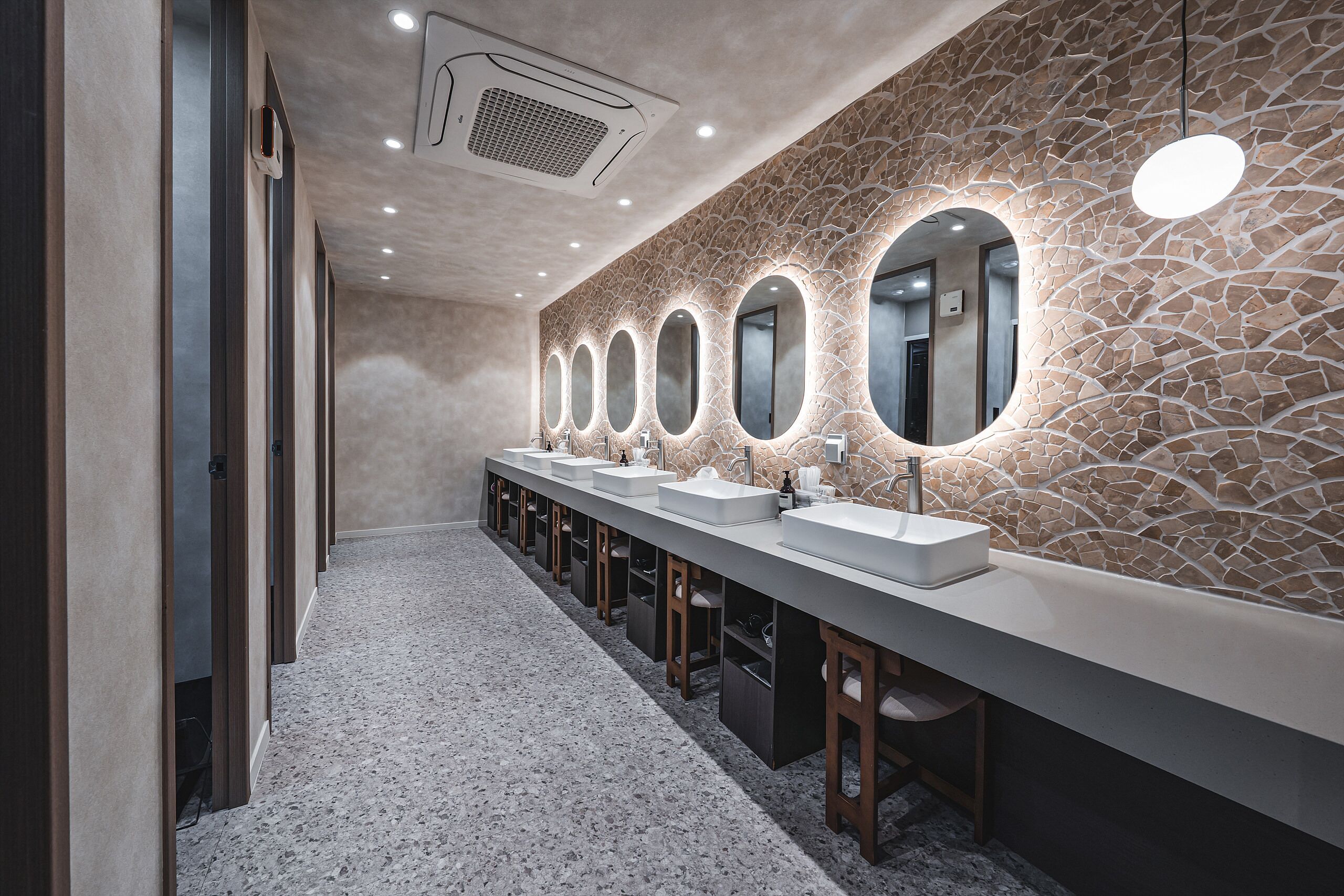 A long, modern bathroom with multiple sinks and mirrors, a stone-patterned wall, and a speckled floor.