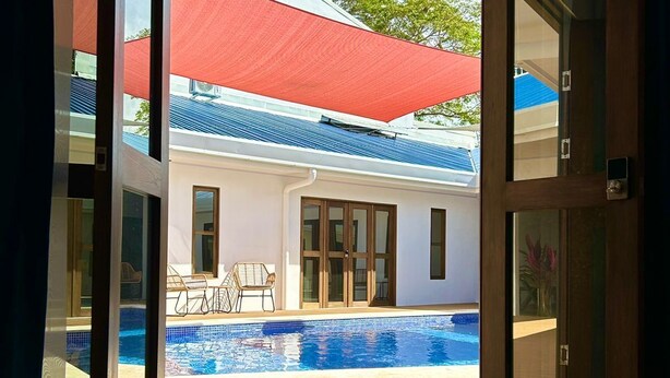 On aperçoit, à travers une porte ouverte, un espace piscine avec une terrasse en bois, un parasol rouge et, au fond, un bâtiment doté d’un toit bleu.