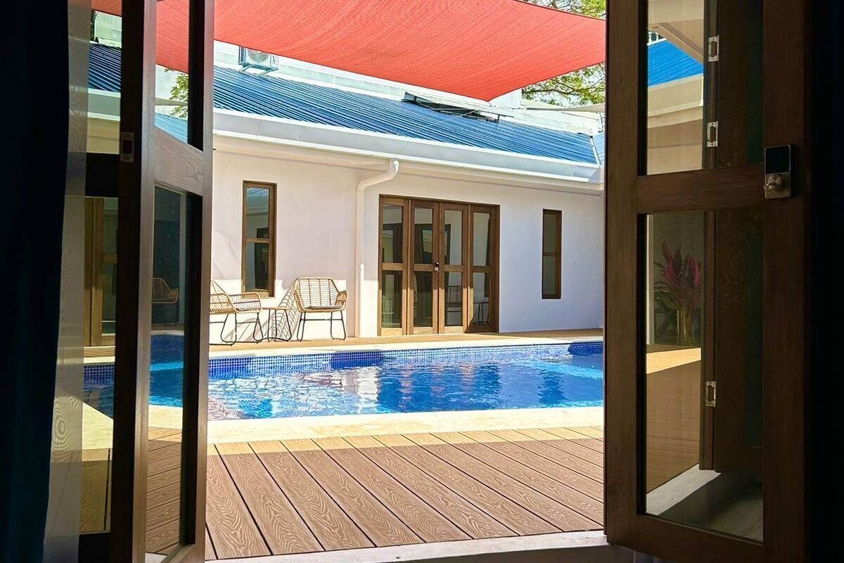 Vista a través de una puerta abierta que da a una zona de piscina con muelle de madera, una sombrilla roja y un edificio con techo azul al fondo.