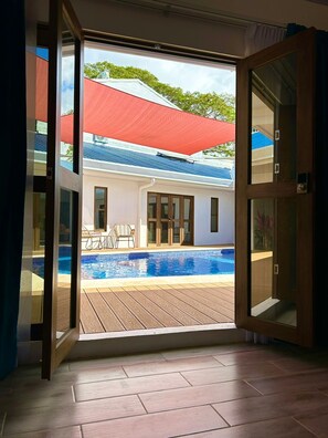 A view through an open door to a swimming pool area with a wooden deck, a red umbrella, and a building with a blue roof in the background. - Hotel Favare (Jaco)