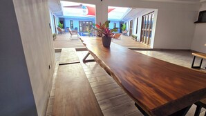Un vaste espace intérieur avec une longue table en bois et des bancs, des plantes en pot et une vue sur un bâtiment bleu avec un toit rouge.