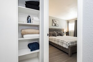 Ein Schlafzimmer mit einem Bett, einem Nachttisch, einem an der Wand befestigten Regal mit gefalteten Handtüchern und einer Tür zu einem anderen Raum.