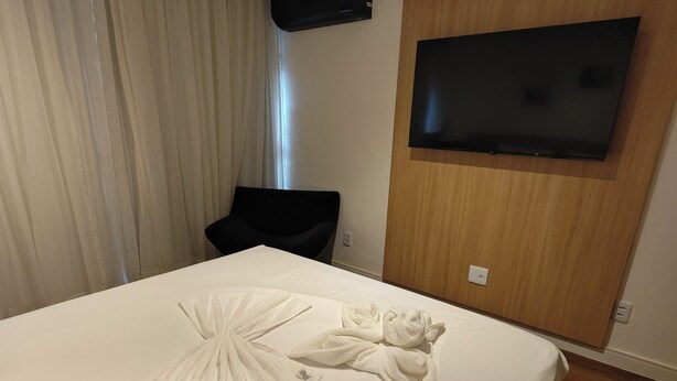 A hotel room with a large bed, a flat-screen TV mounted on the wall, and a wooden panel design. - Flat 109 norte novo e completo (Brasilia)