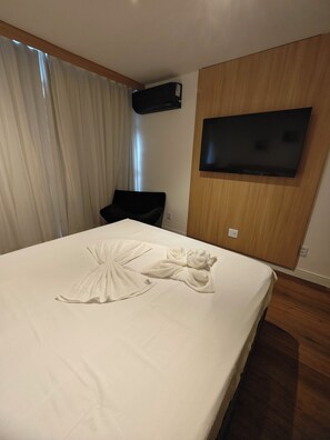 A hotel room with a large bed, a flat-screen TV mounted on the wall, and a wooden panel design. - Flat 109 norte novo e completo (Brasilia)