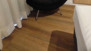 A room with a black chair, a wooden floor, a white bed, and white curtains. - Flat 109 norte novo e completo (Brasilia)
