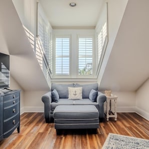 A modern loft bedroom with a sloped ceiling, a grey sofa, a matching ottoman, a small white side table, and a wooden floor.